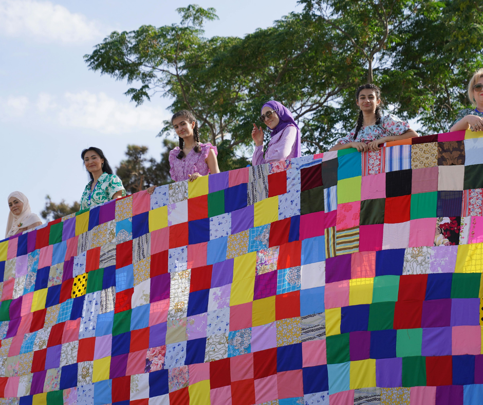 Website Value Collaboration A group of women hold up a complex quilt.